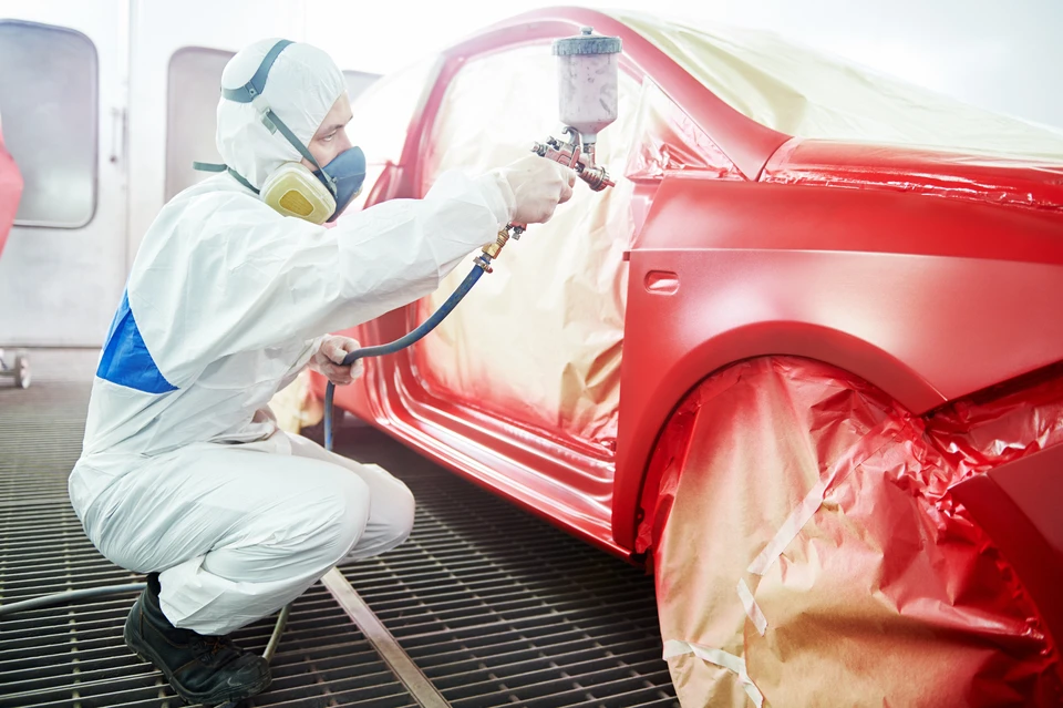 Atelier de carrosserie automobile intérieur avec technicien en combinaison de protection blanche et masque respiratoire appliquant peinture rouge sur véhicule