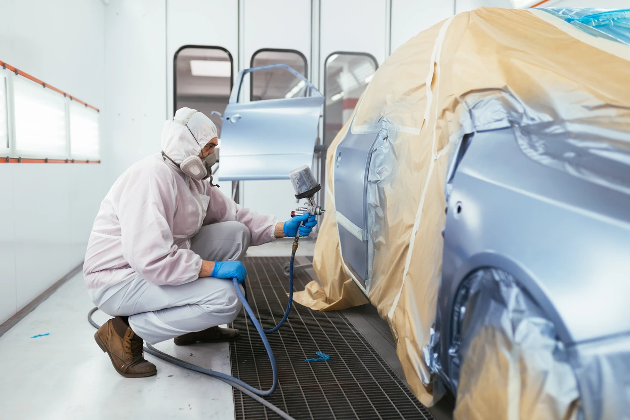 Atelier de carrosserie automobile avec technicien en combinaison de protection intégrale appliquant de la peinture au pistolet sur véhicule