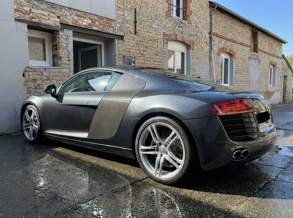 Supercar Audi R8 V8 première génération, coloris gris anthracite métallisé, stationnée devant un bâtiment en pierre