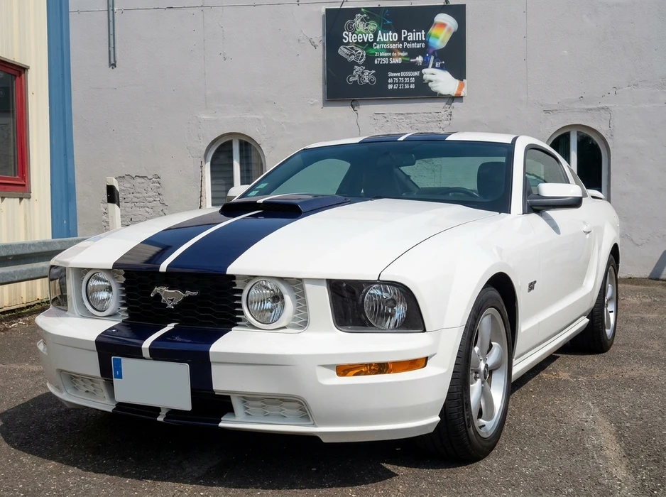 Ford Mustang GT coupé, blanc avec bandes racing bleu marine et blanc, stationnée devant un bâtiment