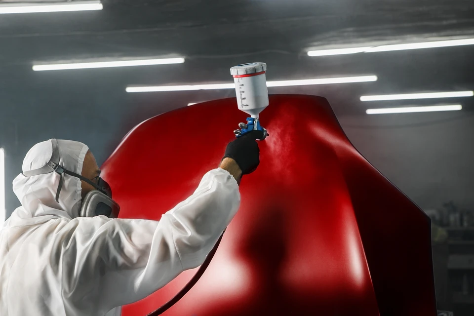Carrossier en combinaison professionnelle travaillant à la préparation ou au ponçage de la carrosserie d'une voiture dans un atelier de carrosserie-peinture, mettant l'accent sur l'expertise technique du spécialiste dans son domaine de compétence (carrosserie et peinture, non mécanique).