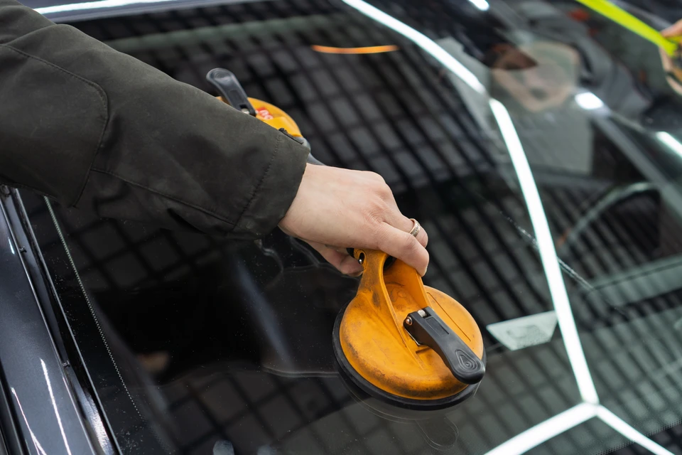 Un technicien en combinaison grise et gants noirs utilise des ventouses jaunes pour remplacer un pare-brise automobile dans un atelier de réparation
